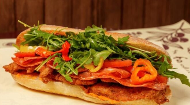 A close-up shot of a large, crusty Italian sub sandwich filled with breaded cutlets, thinly sliced salami, fresh arugula, red and yellow bell pepper slices, and a creamy sauce, resting on a white surface with a patterned napkin in the background.