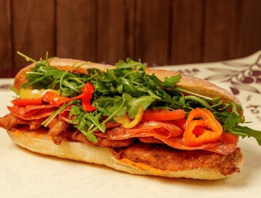 A close-up shot of a large, crusty Italian sub sandwich filled with breaded cutlets, thinly sliced salami, fresh arugula, red and yellow bell pepper slices, and a creamy sauce, resting on a white surface with a patterned napkin in the background.