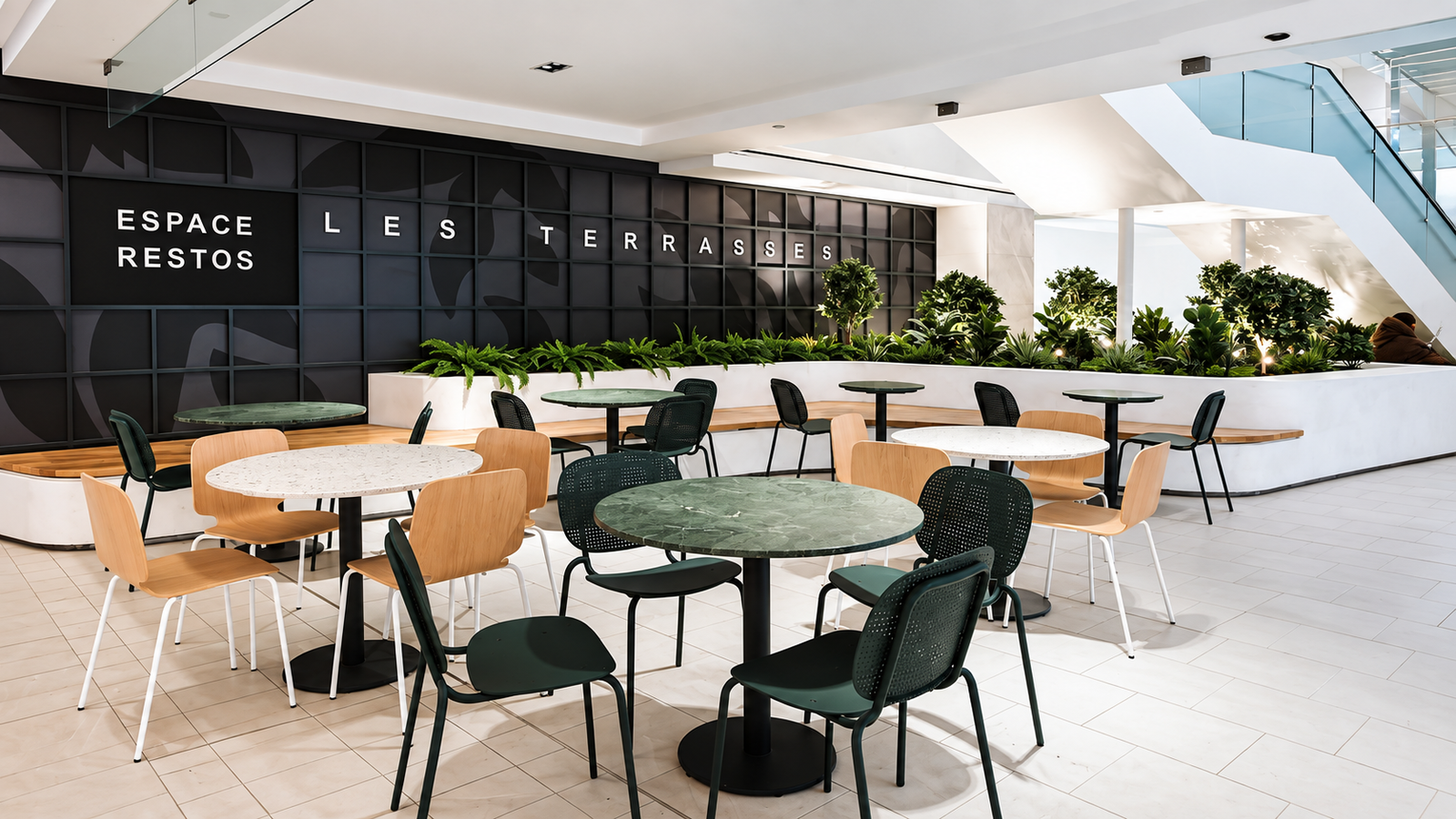A modern, brightly lit indoor dining area features a long black wall with a grid pattern and subtle leaf designs. White letters on the wall read 'ESPACE RESTOS' and 'LES TERRASSES,' above a white curved planter box filled with lush green plants and small trees, which also has an integrated wooden bench. Numerous tables and chairs are arranged on a light tiled floor, with tables having green marble or white speckled tops, and chairs in dark green, light wood, or black; a person is partially visible sitting on the wooden bench in the far background, using a laptop near a white escalator with glass railings.