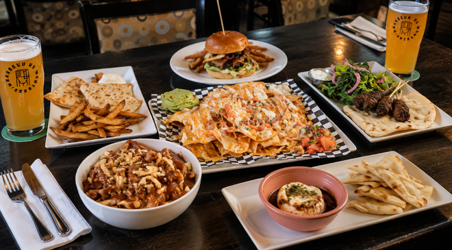 A dark wooden table is covered with a generous spread of pub food and drinks. Prominently featured are a large plate of cheesy nachos with guacamole and salsa, a plate of quesadillas with golden fries, and a bowl of chili cheese fries. Other dishes include a burger with fries, meat skewers with flatbread and a side salad, and a baked cheese appetizer served with pita bread, all accompanied by two glasses of amber beer.