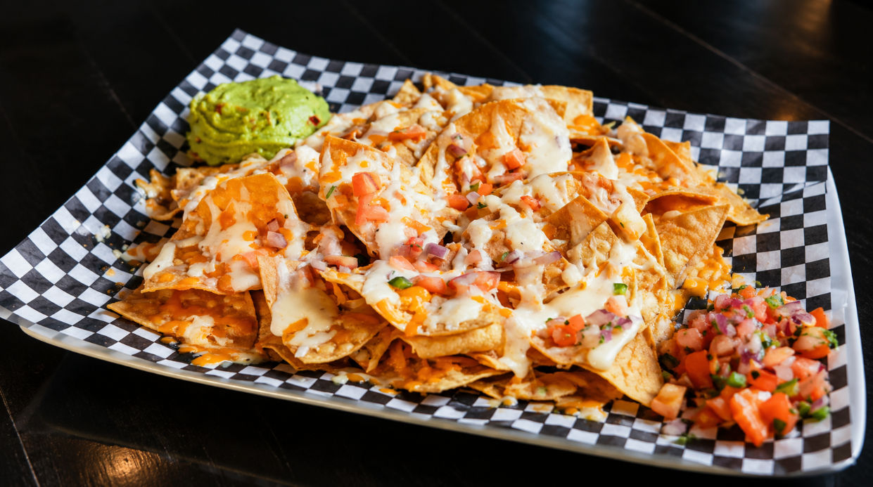 generous spread of pub food and drinks. Prominently featured are a large plate of cheesy nachos with guacamole and salsa, a plate of quesadillas with golden fries, and a bowl of chili cheese fries. Other dishes include a burger with fries, meat skewers with flatbread and a side salad, and a baked cheese appetizer served with pita bread, all accompanied by two glasses of amber beer.