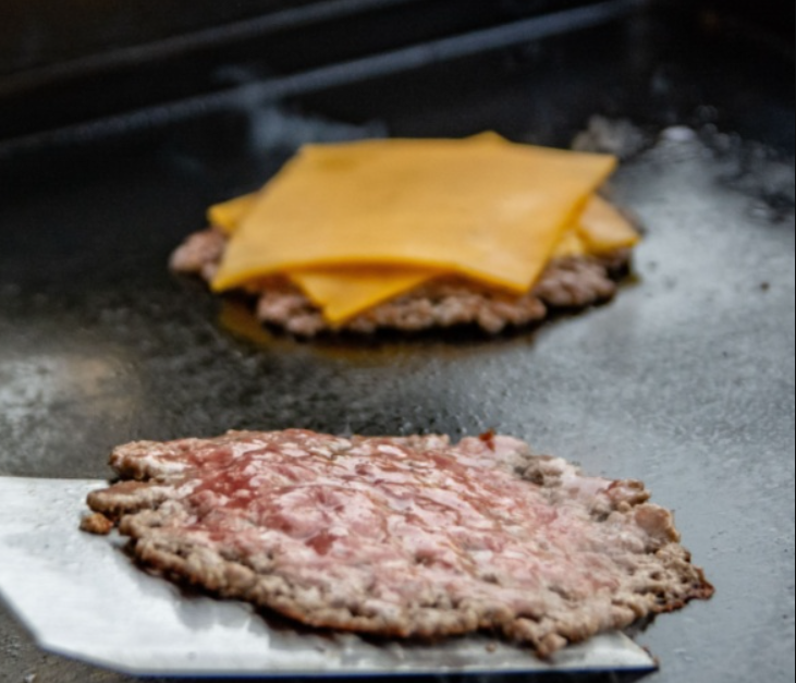 A close-up shot of a smash burger with melted cheese and pickles, partially wrapped in yellow paper, resting on black and white checkered paper. In the background, another burger and a basket of cheese fries are visible, suggesting a fast-food meal setting.
