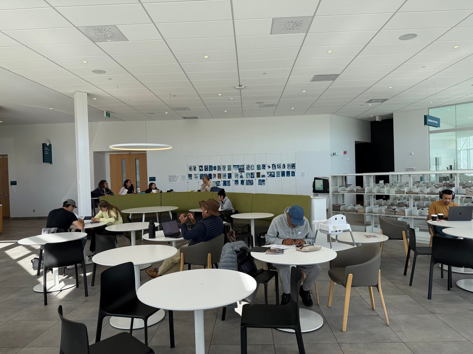 A spacious, brightly lit modern library or study lounge features people seated at round white tables working on laptops and reading. The room has light wood-look flooring, white ceiling panels with recessed lighting, green booth seating along the back wall, and shelves filled with books to the right.
