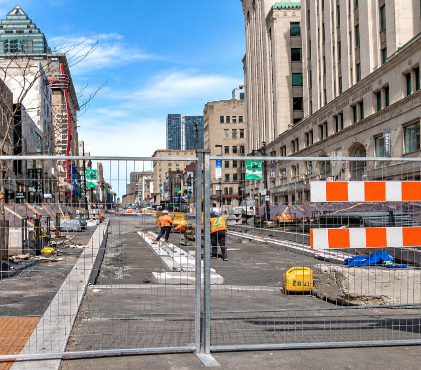 An outdoor city street scene showing construction barriers with orange and white striped wooden rails. Two orange signs with black text in French read "RUE BARRÉE" (street closed) and "DÉTOUR" (detour) with an arrow indicating a U-turn. Blue temporary fencing, snow piles at the base, and historic European-style architecture are visible in the background under a blue sky with scattered clouds.