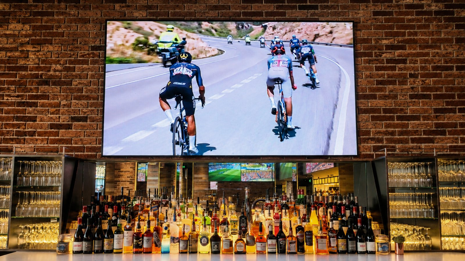 A large screen mounted on a brick wall displays a cycling race with several cyclists riding down a winding road, positioned above a heavily stocked bar with rows of glasses and liquor bottles on display.