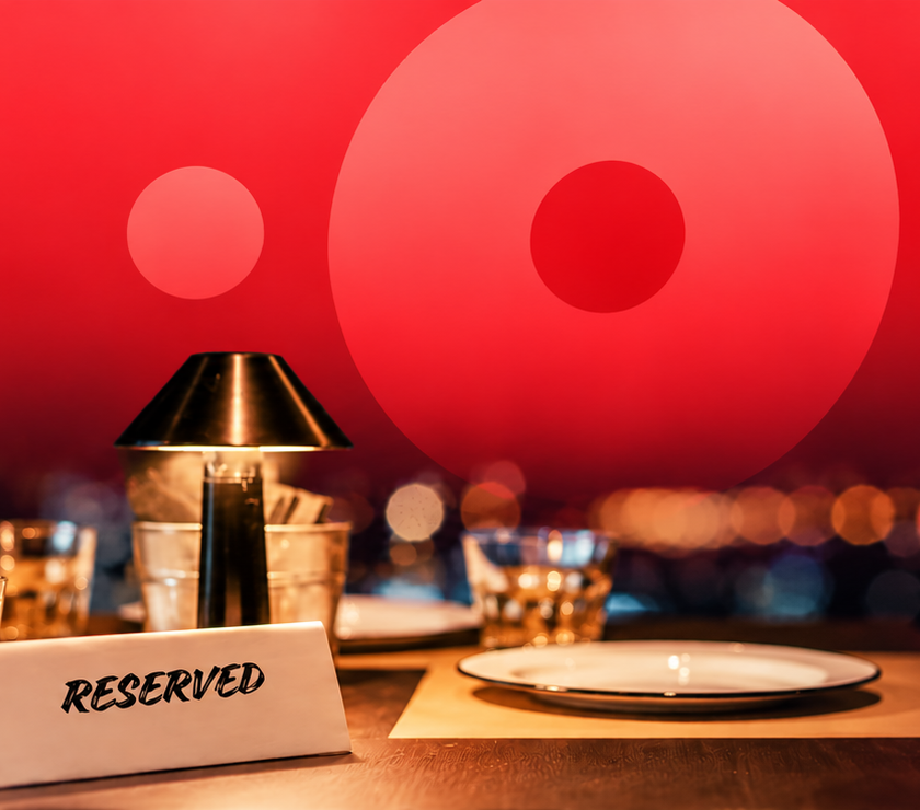 A close-up view of a restaurant table set for dinner with a 'RESERVED' sign, a small lamp, glasses, and a plate, against a blurred backdrop of city lights and a vibrant red sunset.