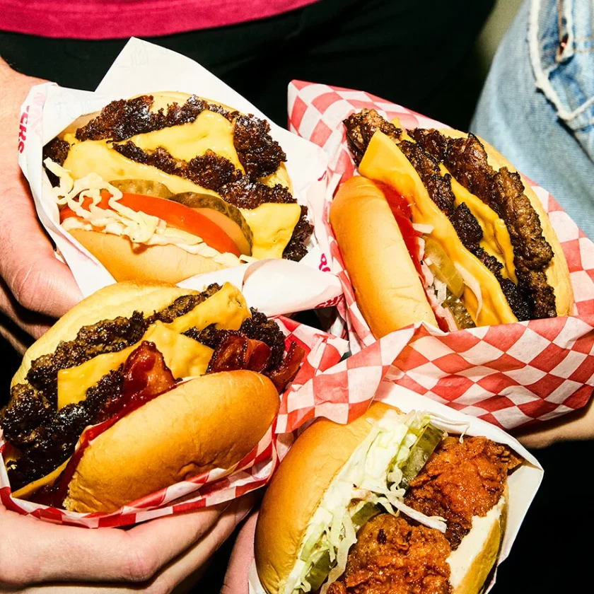 A close-up view of several loaded cheeseburgers served in red and white checkered paper wrappers, held by someone's hands.